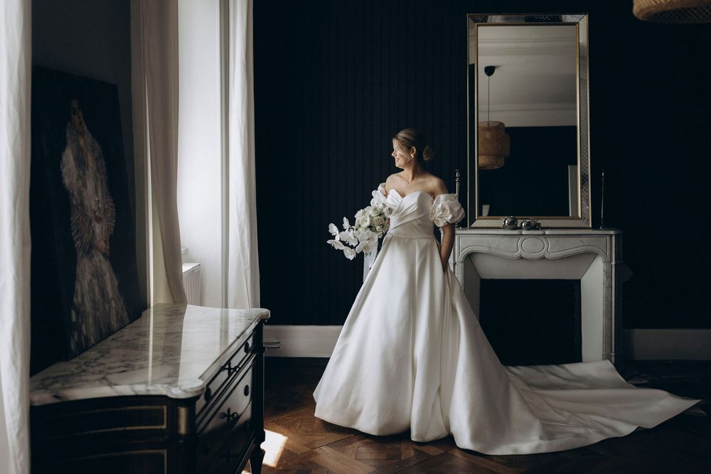 A bride in a flowing white wedding gown stands elegantly in a dark room with natural window light.