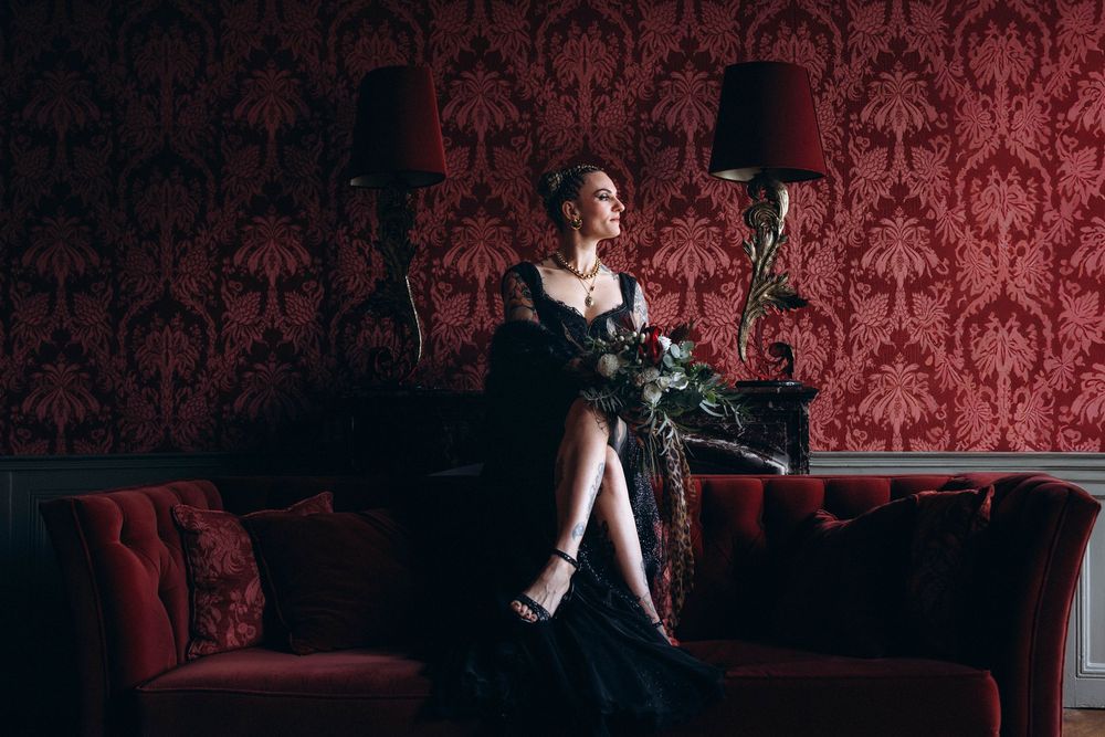 A person in a black dress poses against a red damask wallpaper in a dramatic fashion photography series.
