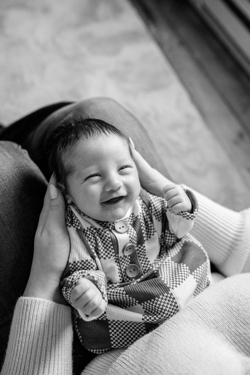 Black and white portrait of a joyful baby wearing a checkered outfit, smiling while being held.
