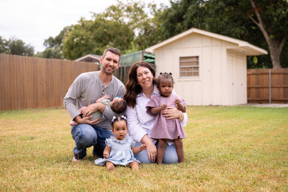 A family sits together on the grass in their backyard with a white house and wooden fence in the background.