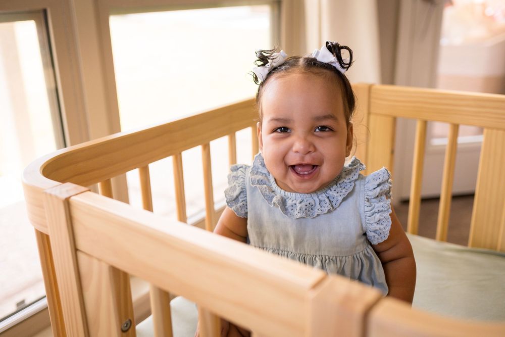 A joyful baby in a light blue outfit sits in a wooden crib while smiling and laughing.