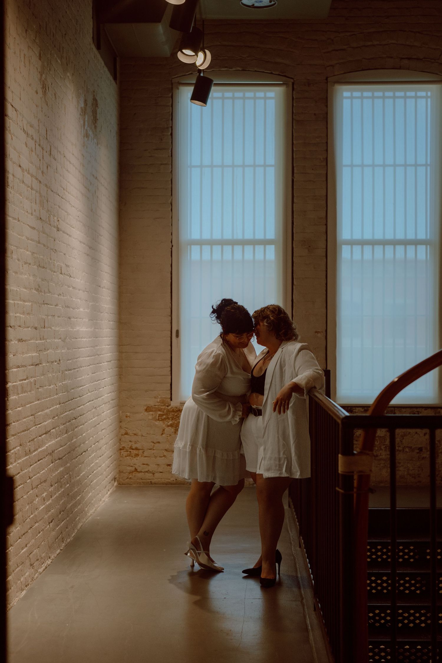 Two people sharing an intimate moment in a sunlit hallway with brick walls and tall windows.