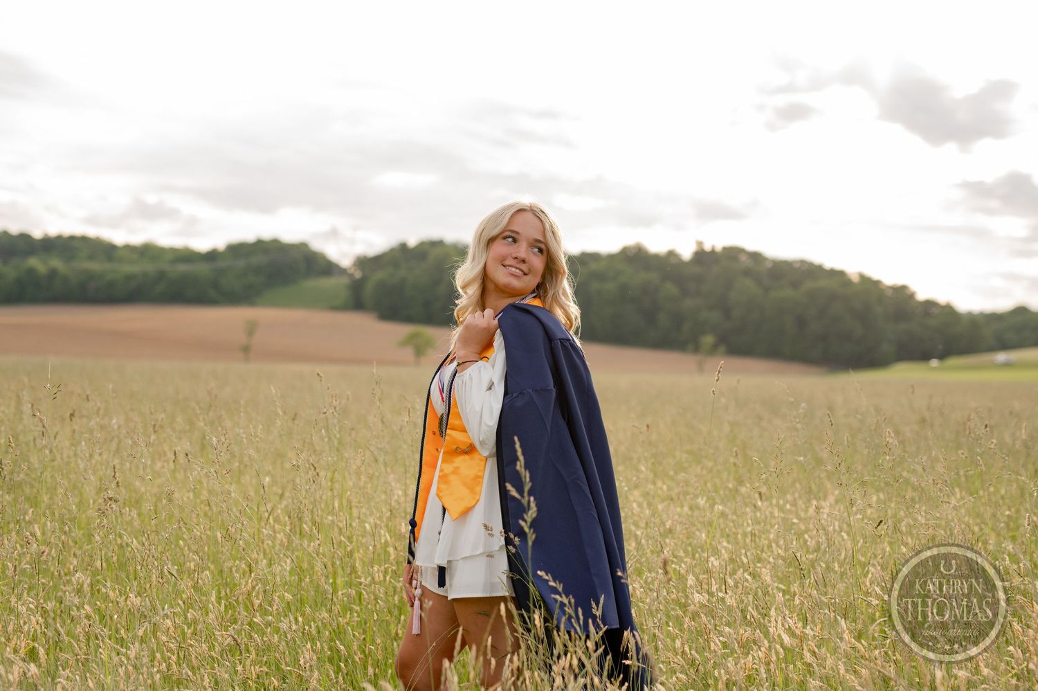 Graduate in academic regalia poses in golden field at sunset.