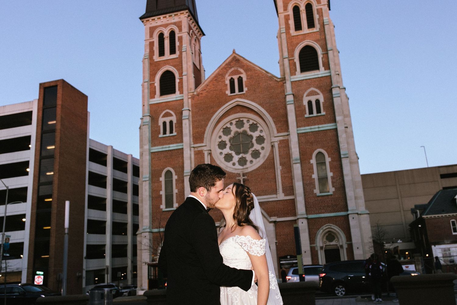 Historic red brick church exterior with twin towers provides backdrop for wedding portrait.
