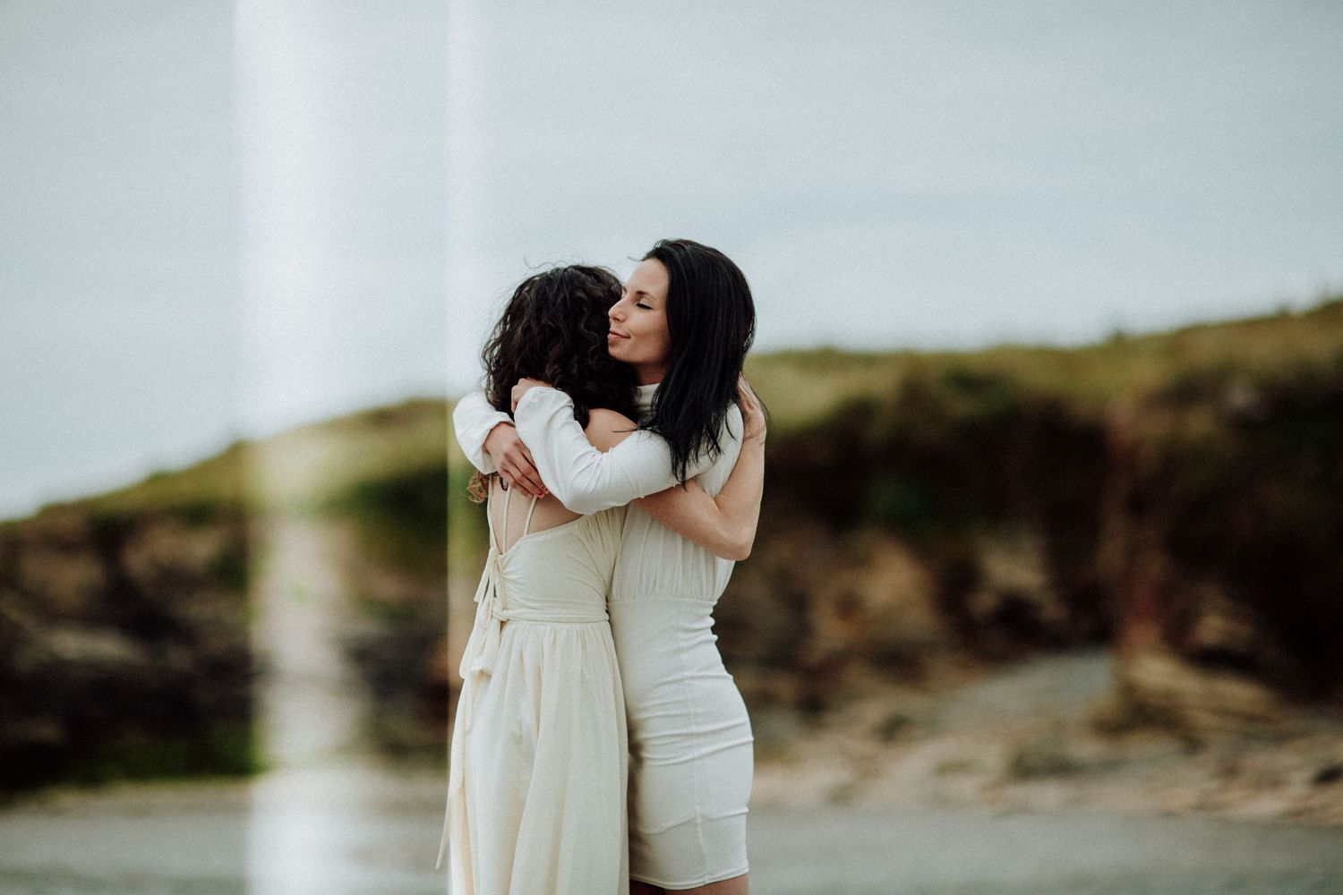 Two people in white dresses sharing an intimate embrace on a rocky coastline.