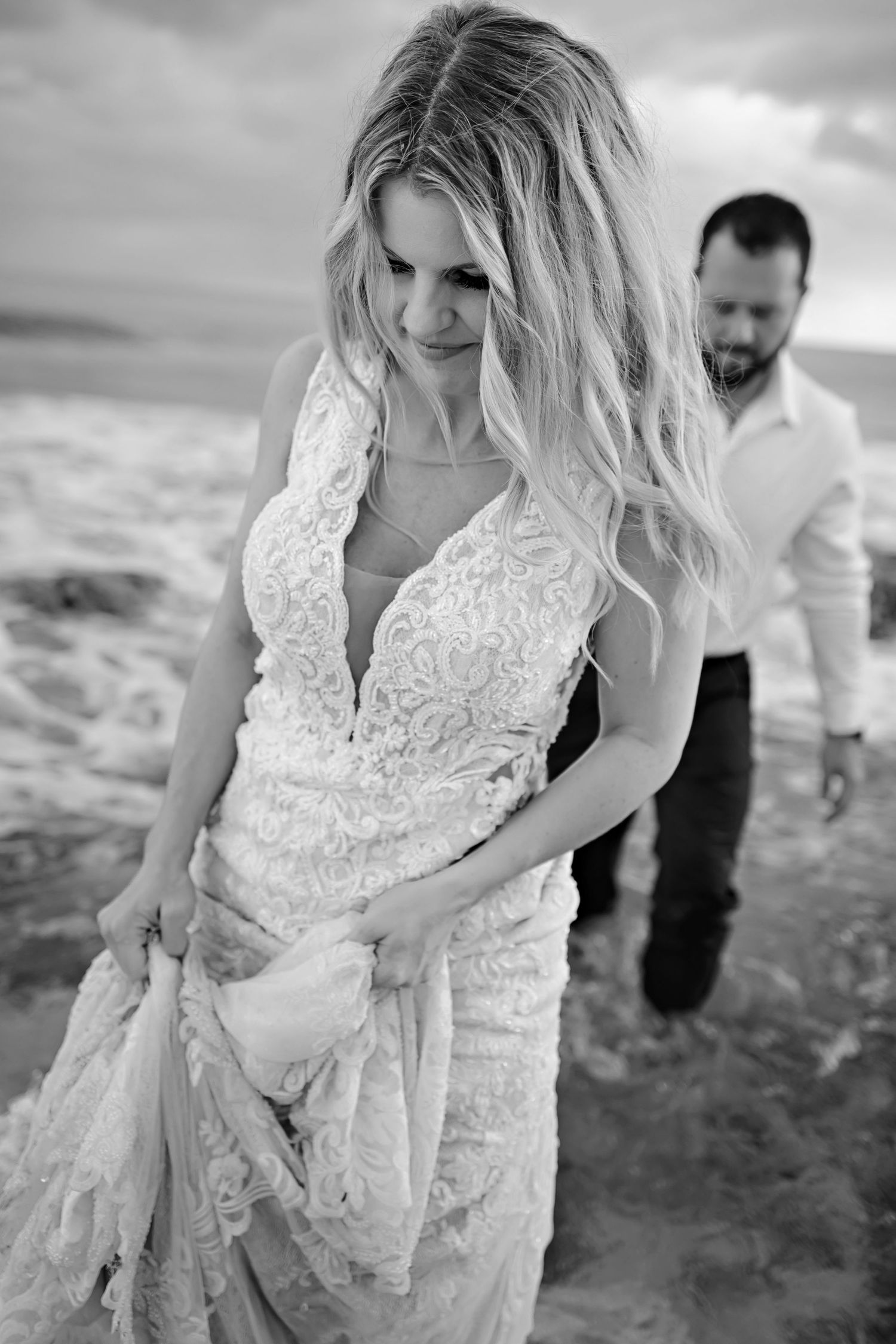 A black and white beach wedding photo showing a bride in a lace dress walking in the water with the groom behind her.