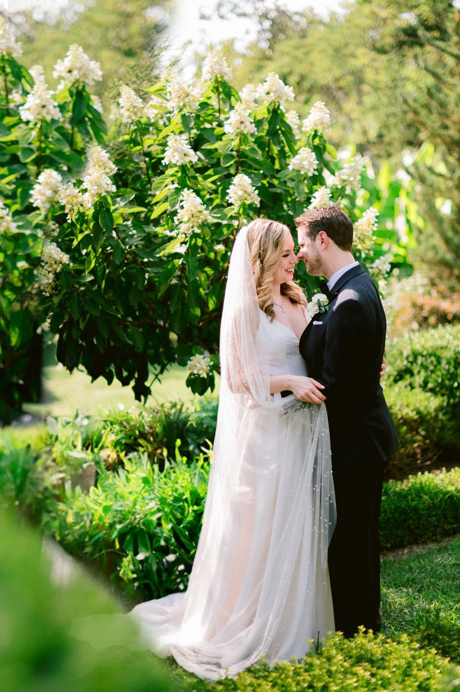 Romantic wedding couple portraits in lush garden setting with blooming white flowers.