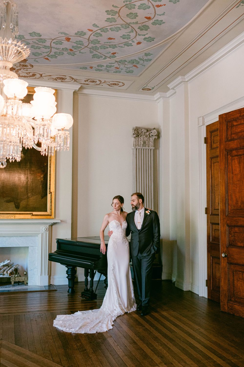 Elegant wedding portrait in historic room with ornate ceiling, chandelier, and grand piano.