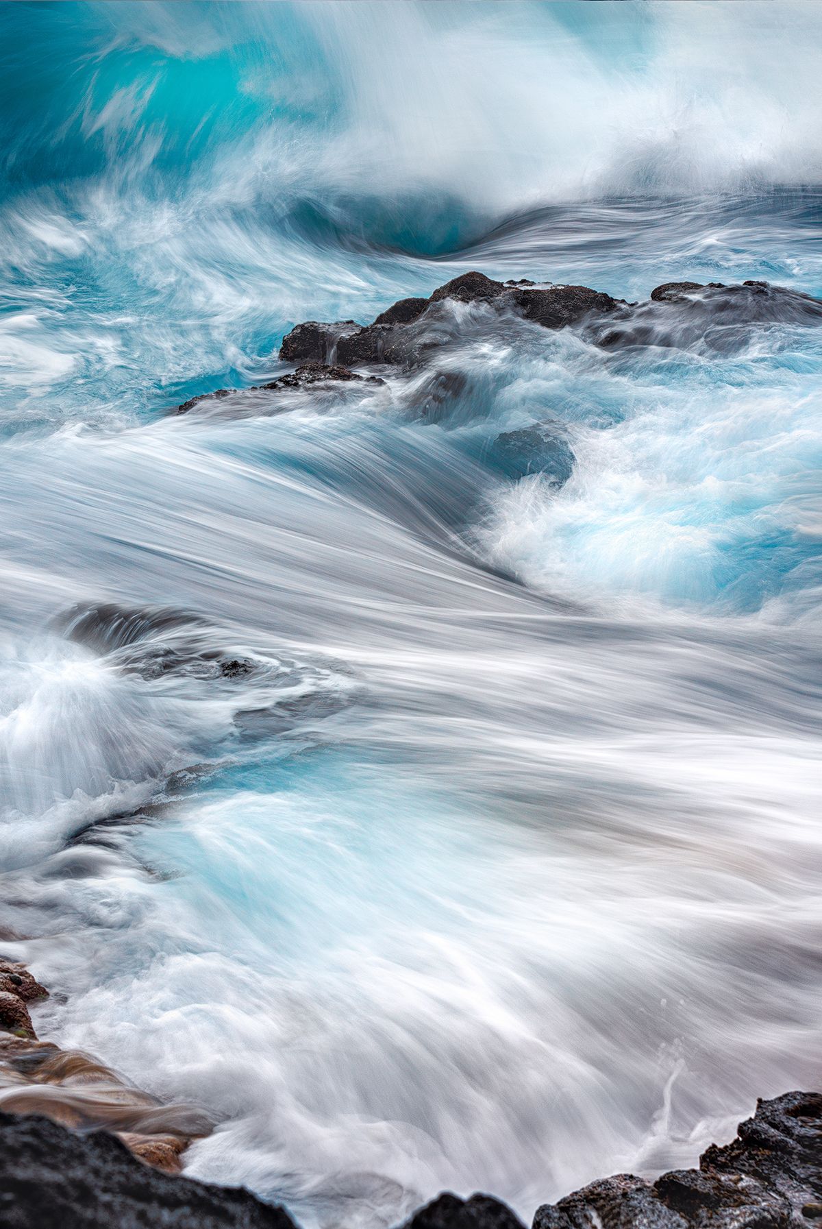 Powerful ocean waves creating a turquoise and white cascade over dark coastal rocks.