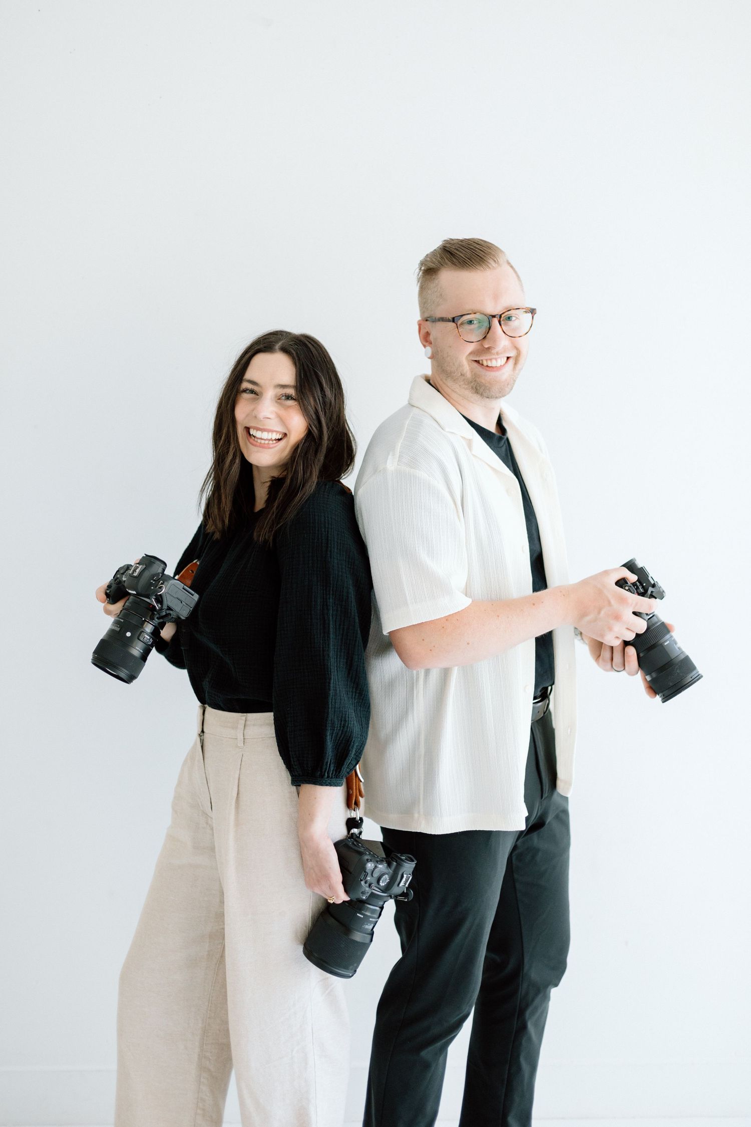 Two photographers stand back to back holding professional cameras in a studio setting.