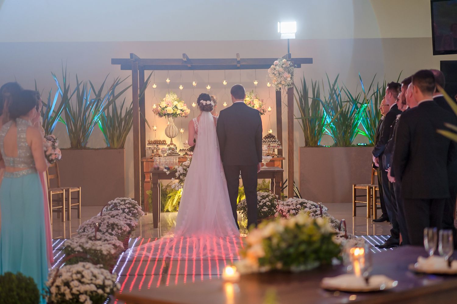 Wedding ceremony view from behind showing couple at altar with guests standing along decorated aisle.