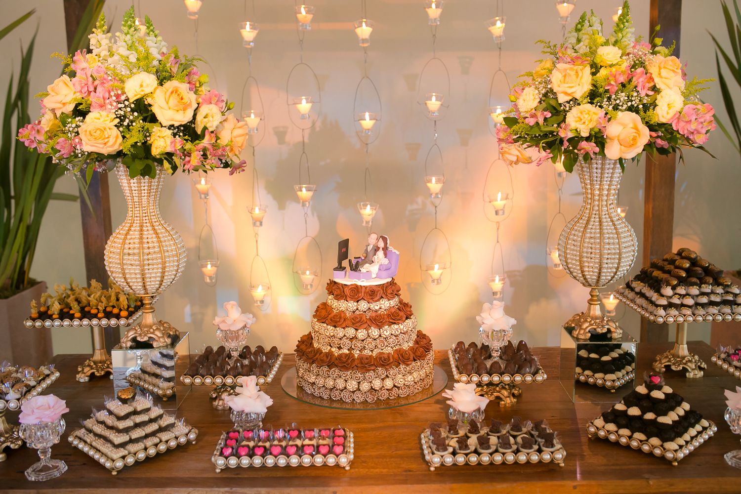 Elegant dessert display table with tiered cake, flowers in white vases, and twinkling string lights creating a romantic ambiance.