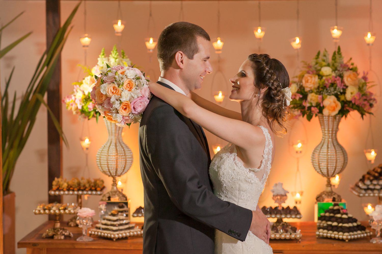 Couple embraces in front of elegant floral arrangements and dessert display at wedding reception.