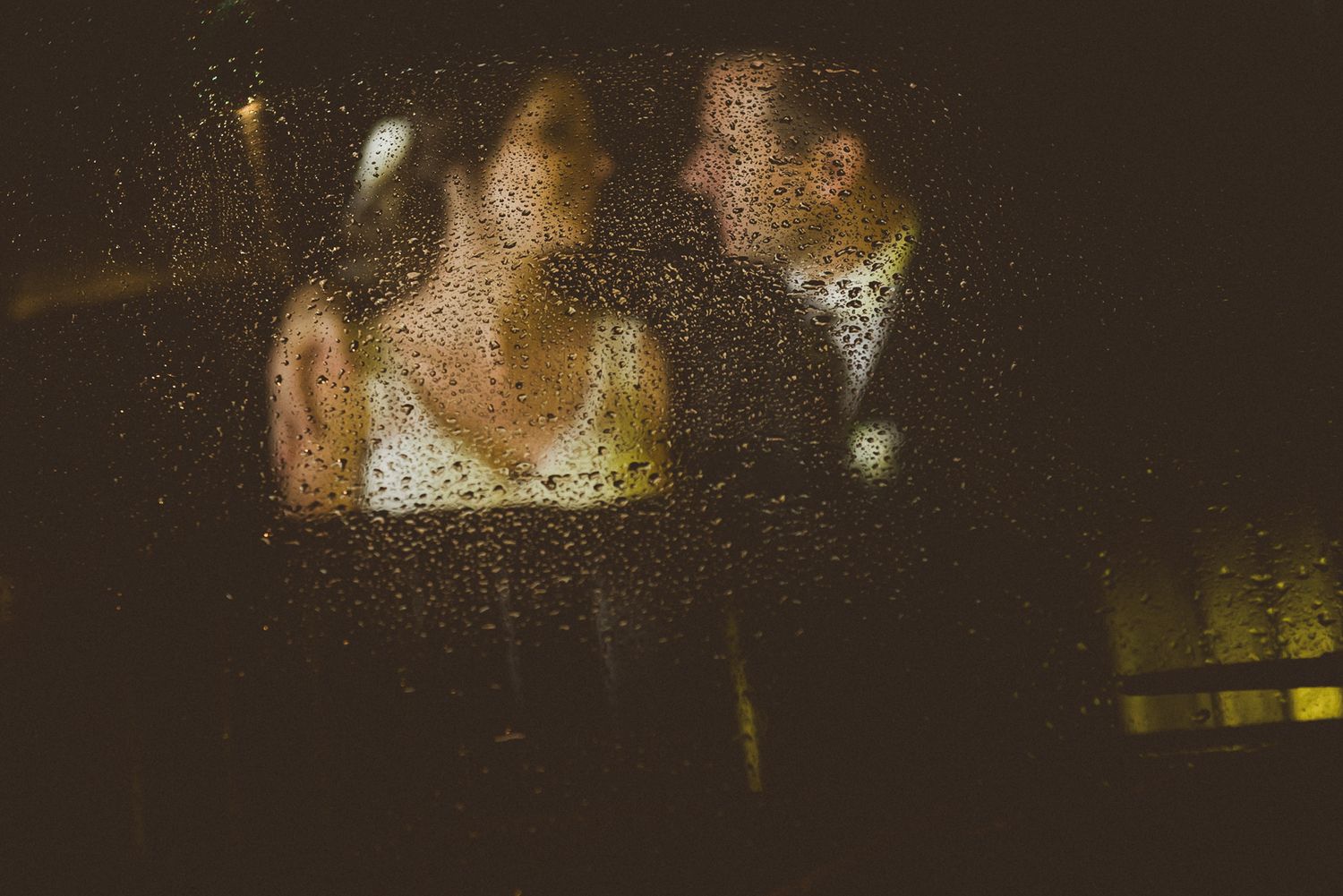 Rain drops on window create romantic bokeh effect around dancing figures at evening celebration.