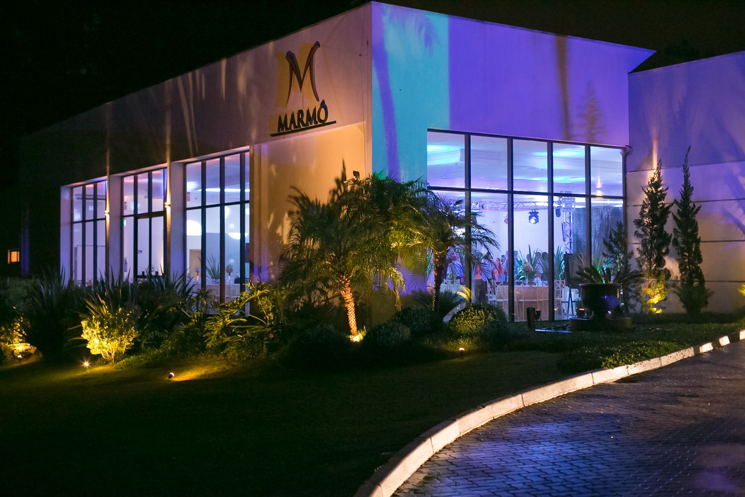 Modern event venue exterior illuminated at night with curved pathway and landscaping.