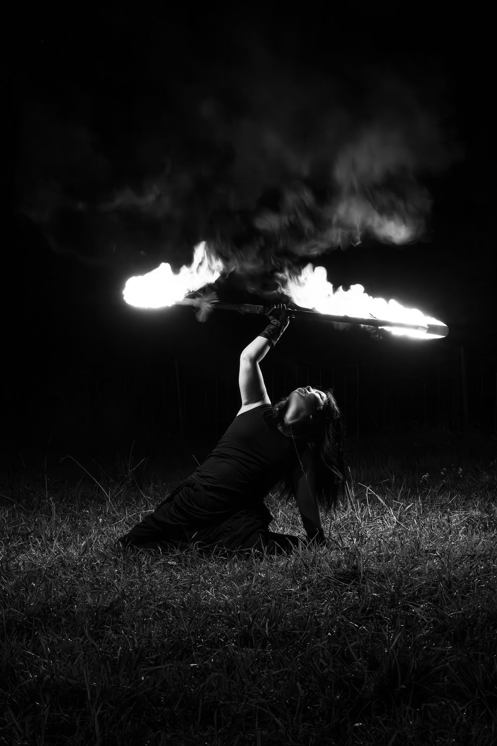 Black and white photo of a woman performing fire dancing outdoors at night, spinning a flaming staff.