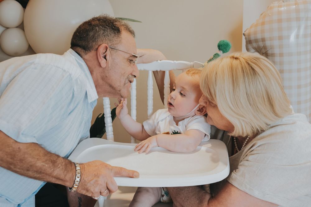 Grandparents lovingly interact with a baby in a high chair at a birthday party with balloon decorations.