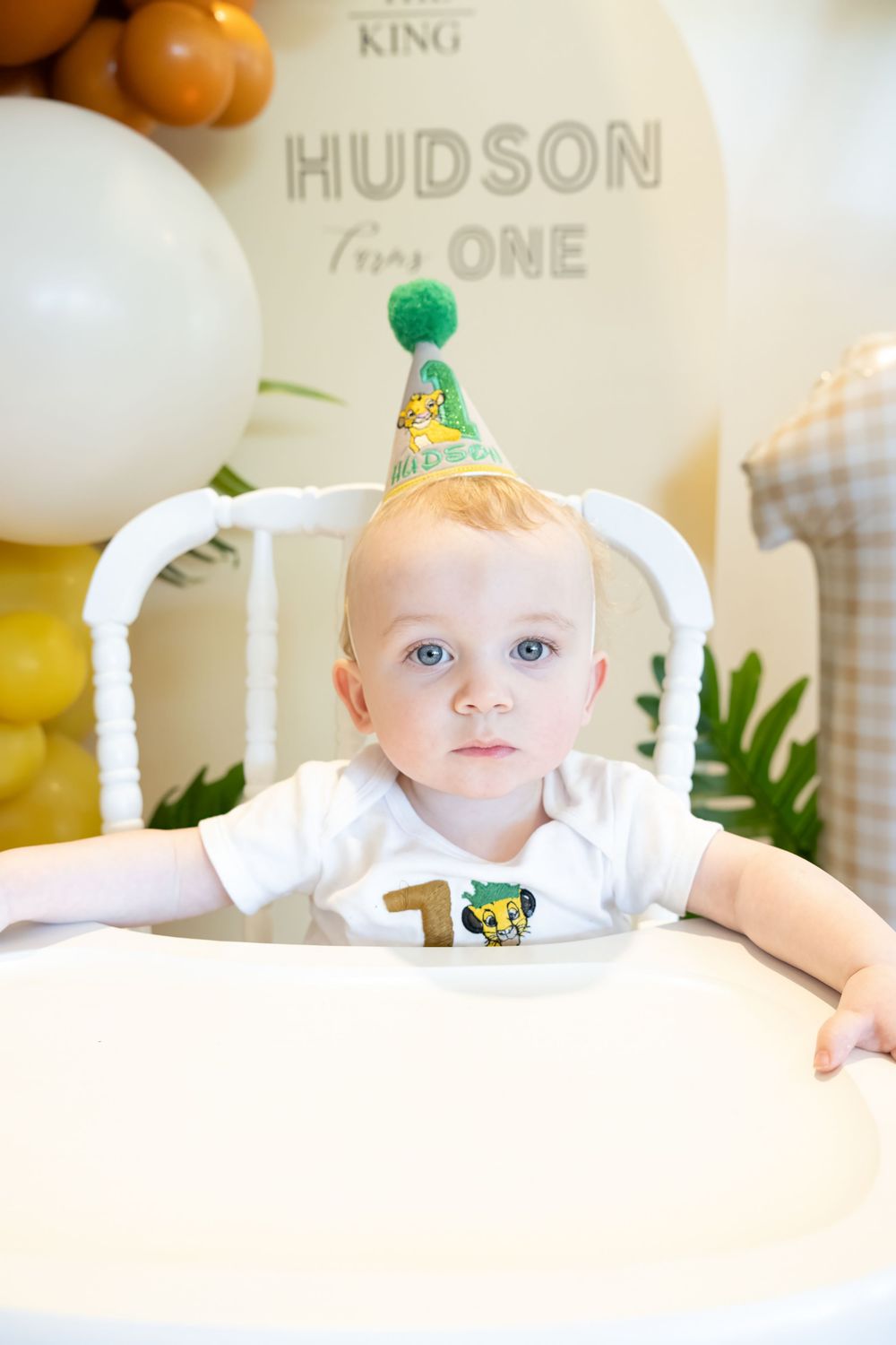 Baby boy Hudson celebrates his first birthday in a high chair wearing a Lion King party hat with number one decorations.