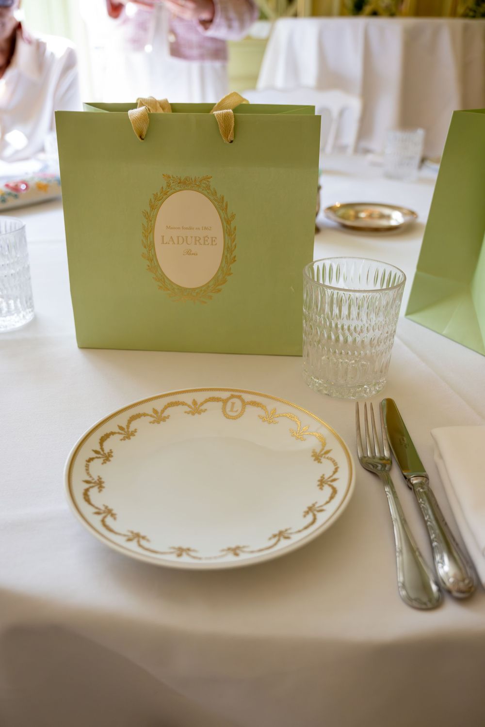 Elegant Ladurée gift bag beside a gold-rimmed white plate, crystal glass, and silver cutlery on a white tablecloth.