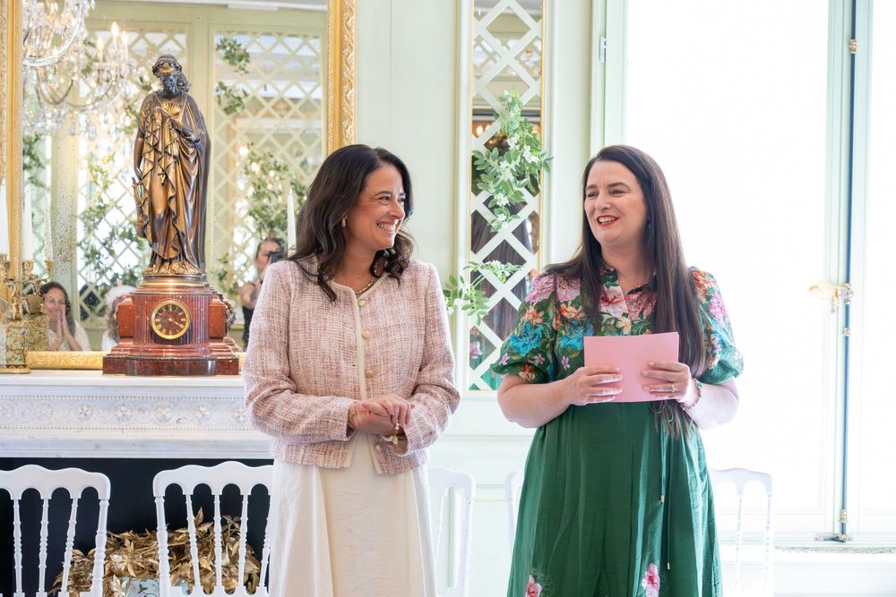 Two elegantly dressed women smile while one holds a pink folder in a ornate white and gold decorated room.