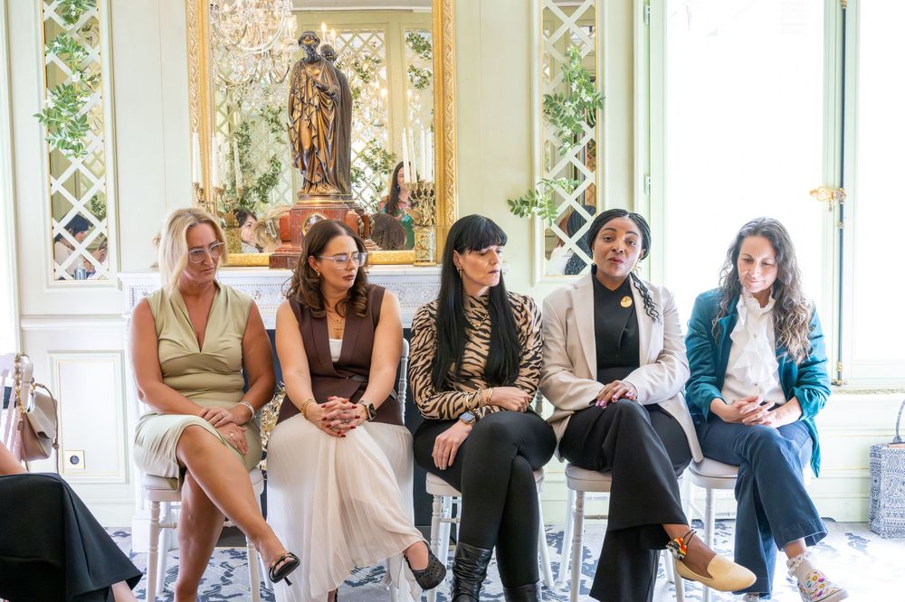 Five women seated in elegant room with ornate decor, engaged in panel discussion at a formal event.