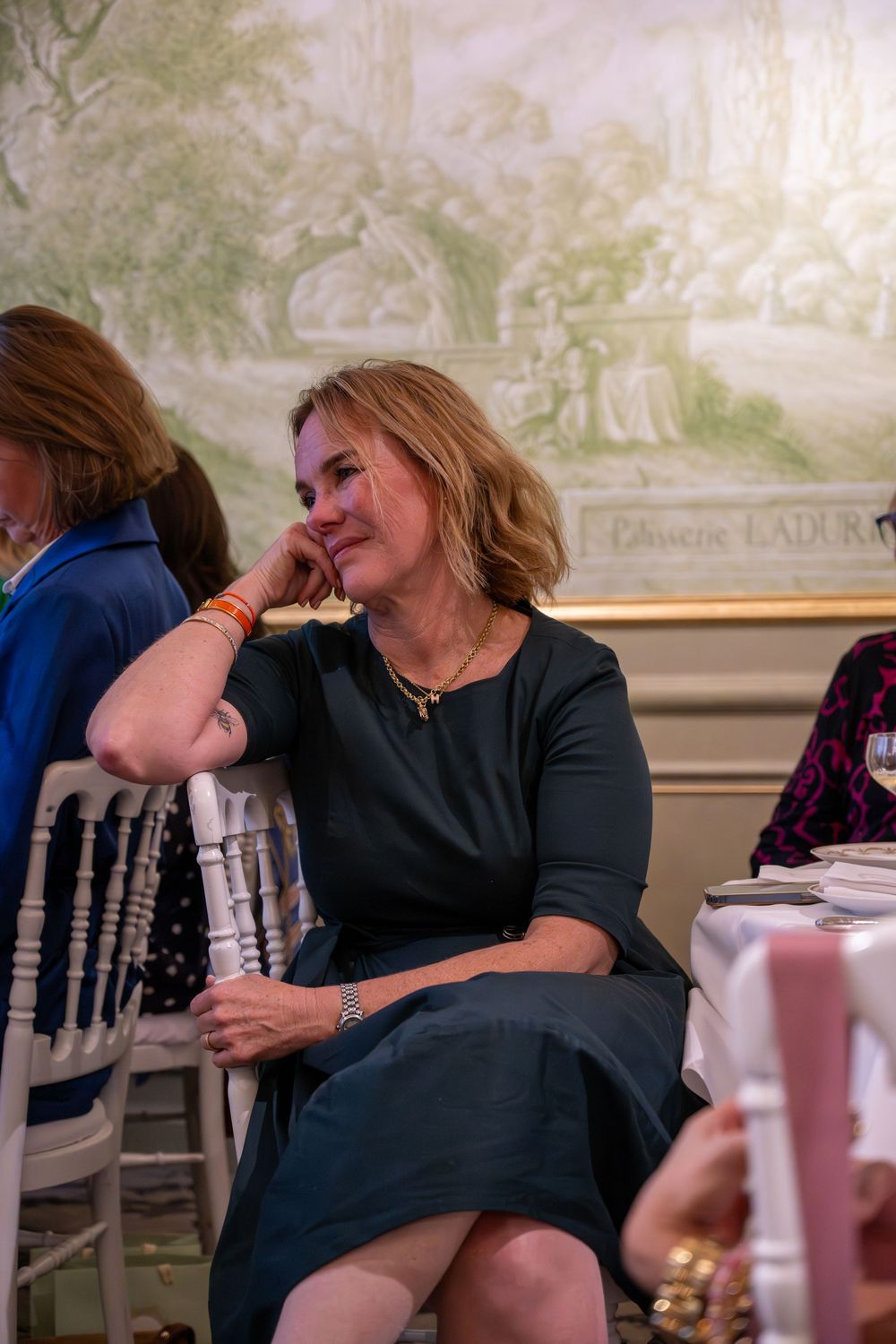Woman in teal dress sits relaxed at elegant event table, smiling thoughtfully with mural backdrop.