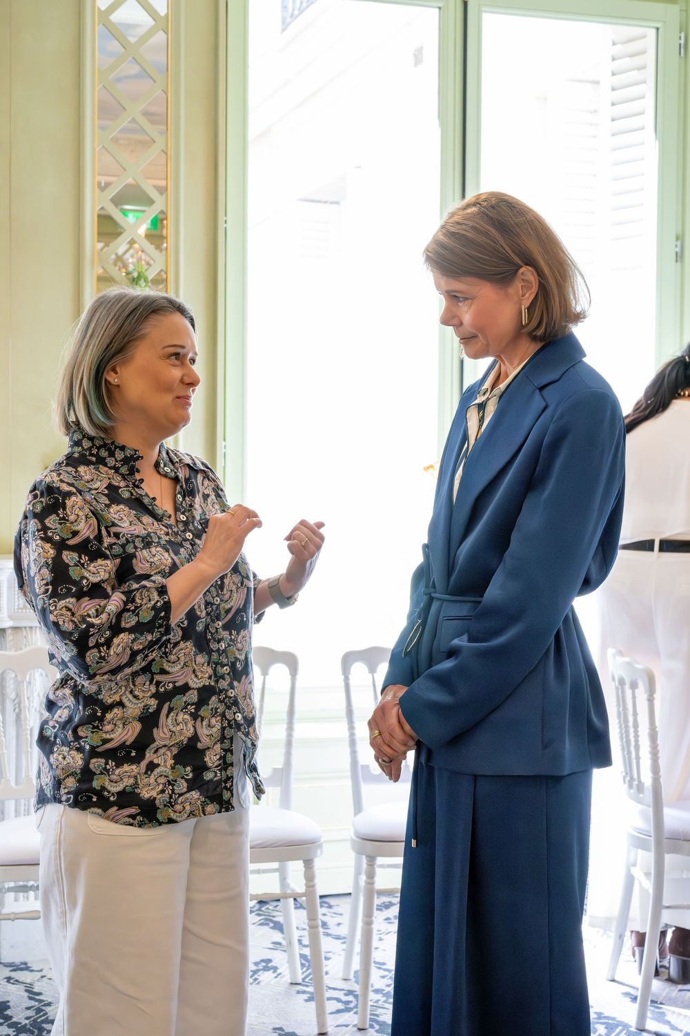 Two women in elegant attire engage in conversation inside a bright, decorated room with white chairs.
