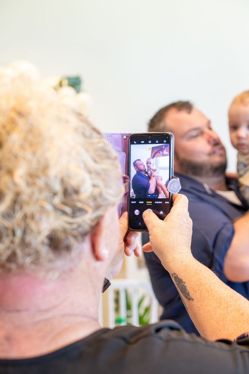 Person with curly blonde hair photographs a man holding a toddler on a smartphone in a bright indoor setting.