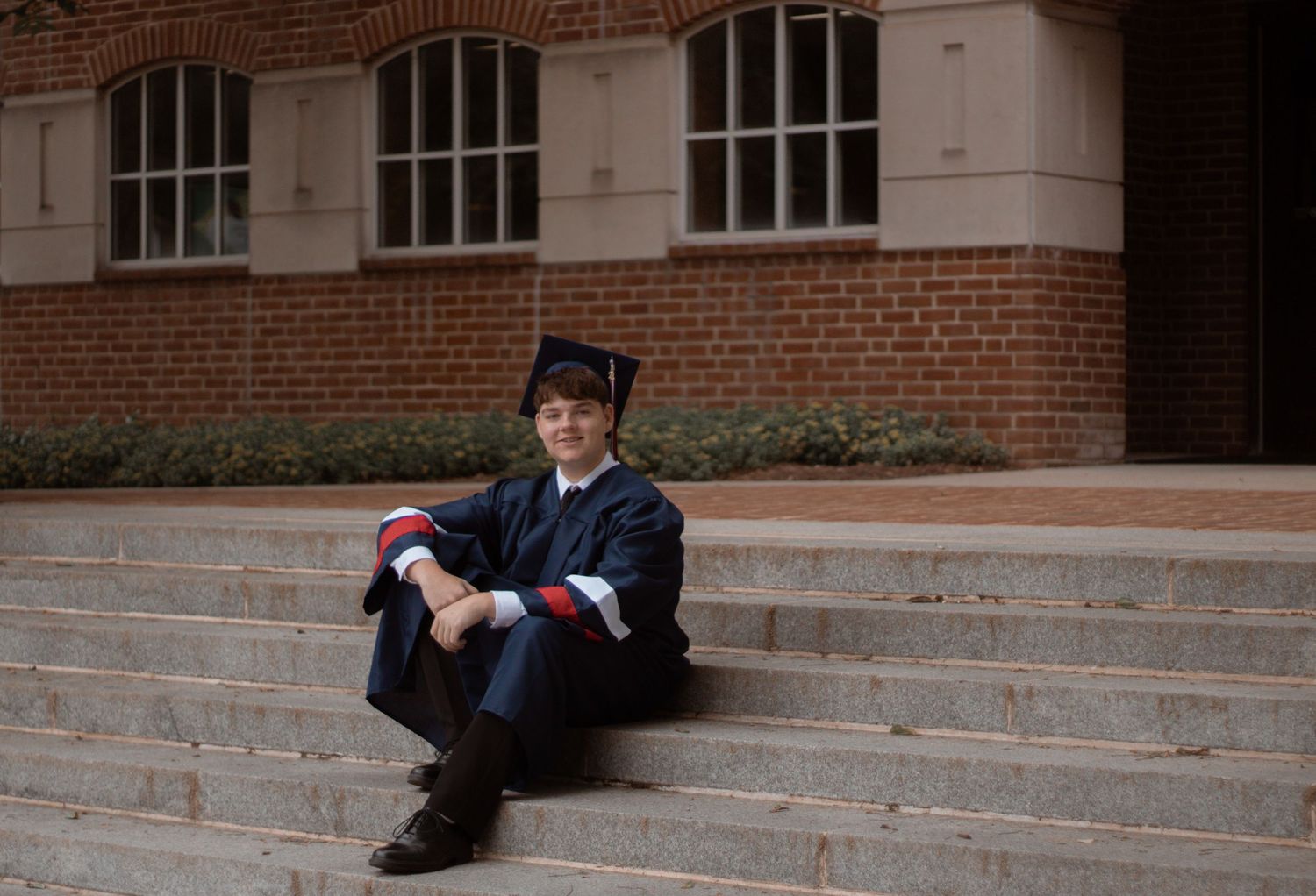 Graduate in black cap and gown sitting on stone steps of a brick university building.