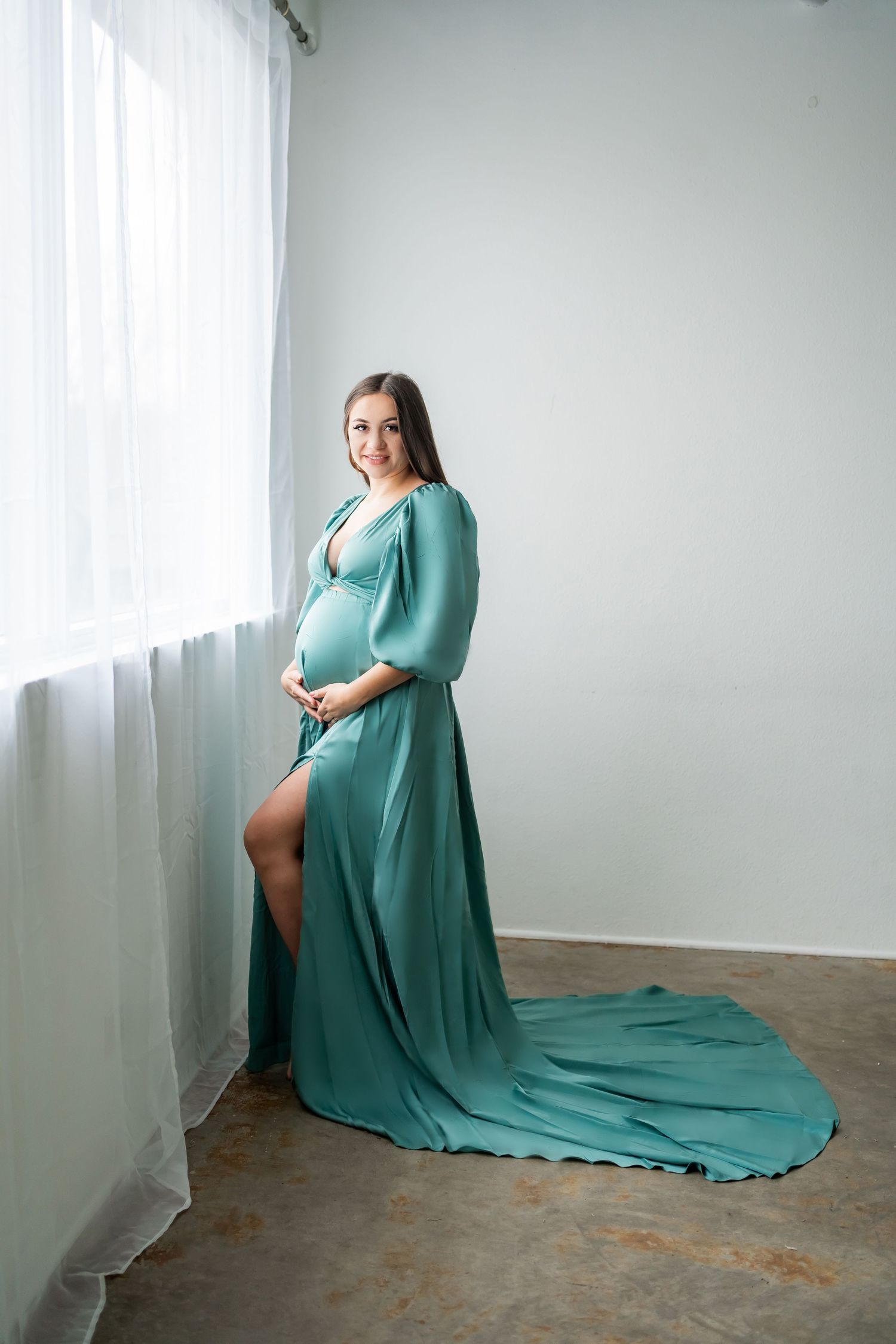 Elegant maternity photo featuring a flowing teal gown with long train against a white studio backdrop.