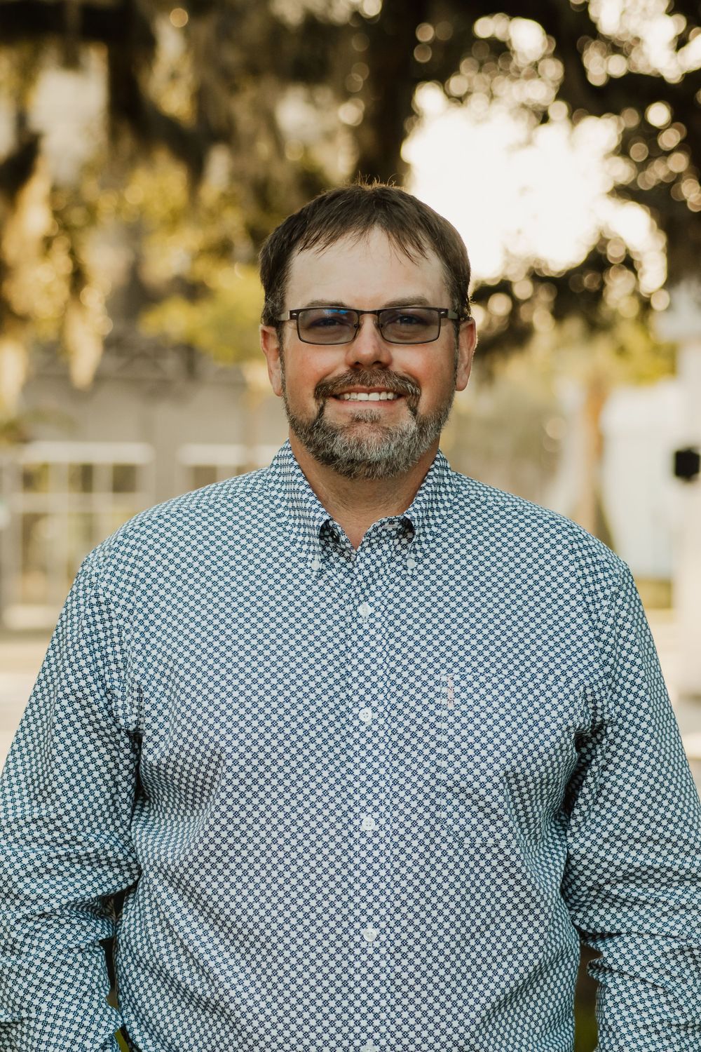 Professional portrait of person wearing glasses and blue checkered shirt outdoors with blurred trees in background.