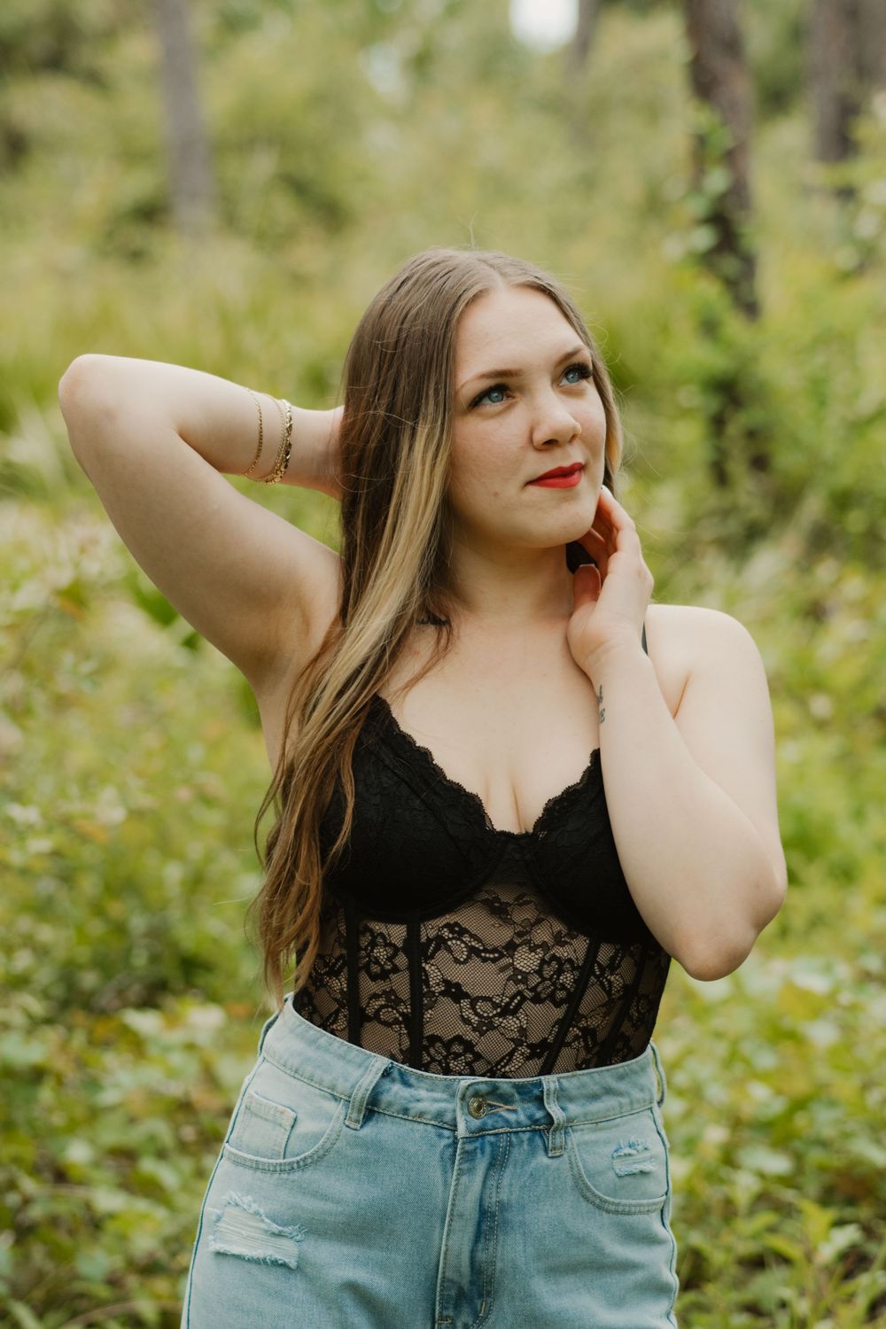 A woman in a black lace top and denim jeans poses in a natural outdoor setting with green foliage.