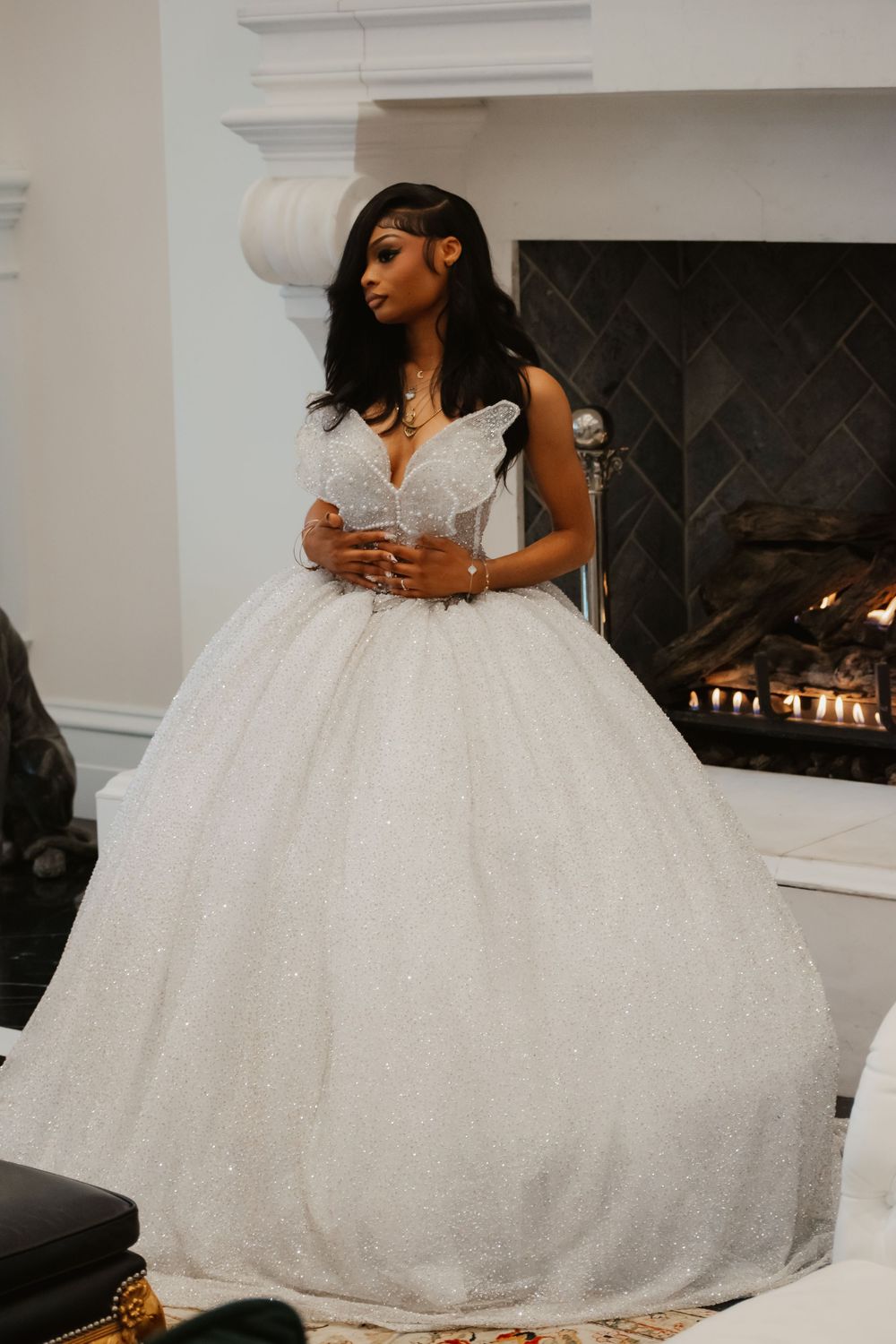 Bride in sparkly white ball gown standing elegantly near a lit fireplace indoors.