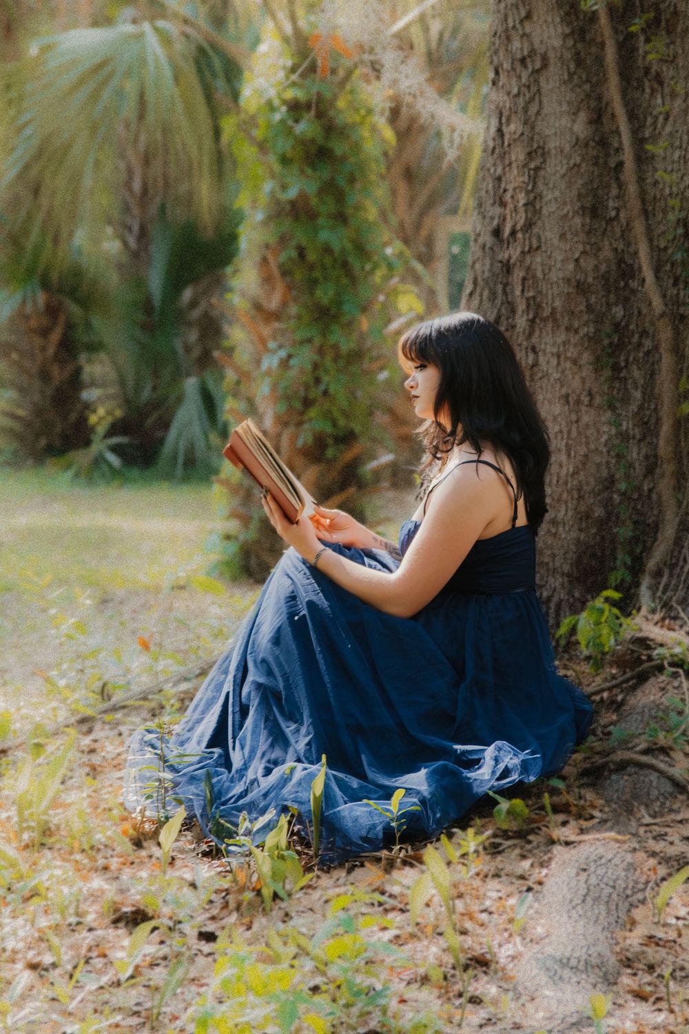 Woman in blue maxi dress reading a book while sitting against a tree in a lush garden.