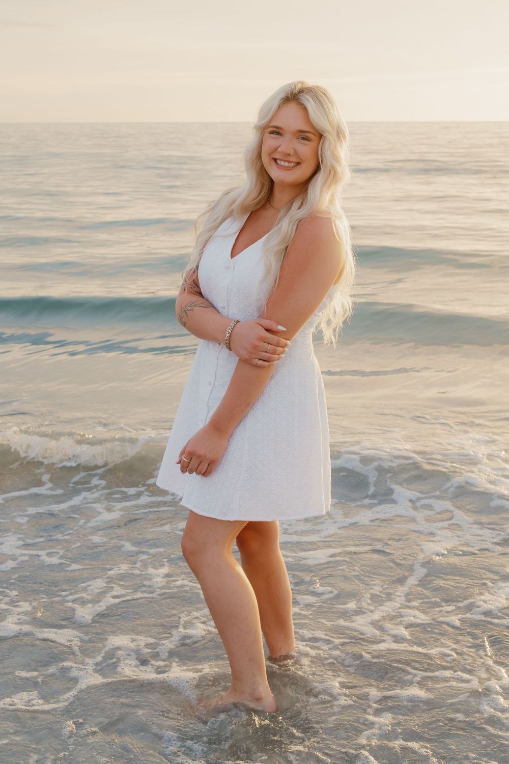 Young blonde woman in white dress standing barefoot in ocean waves at sunset on a sandy beach.