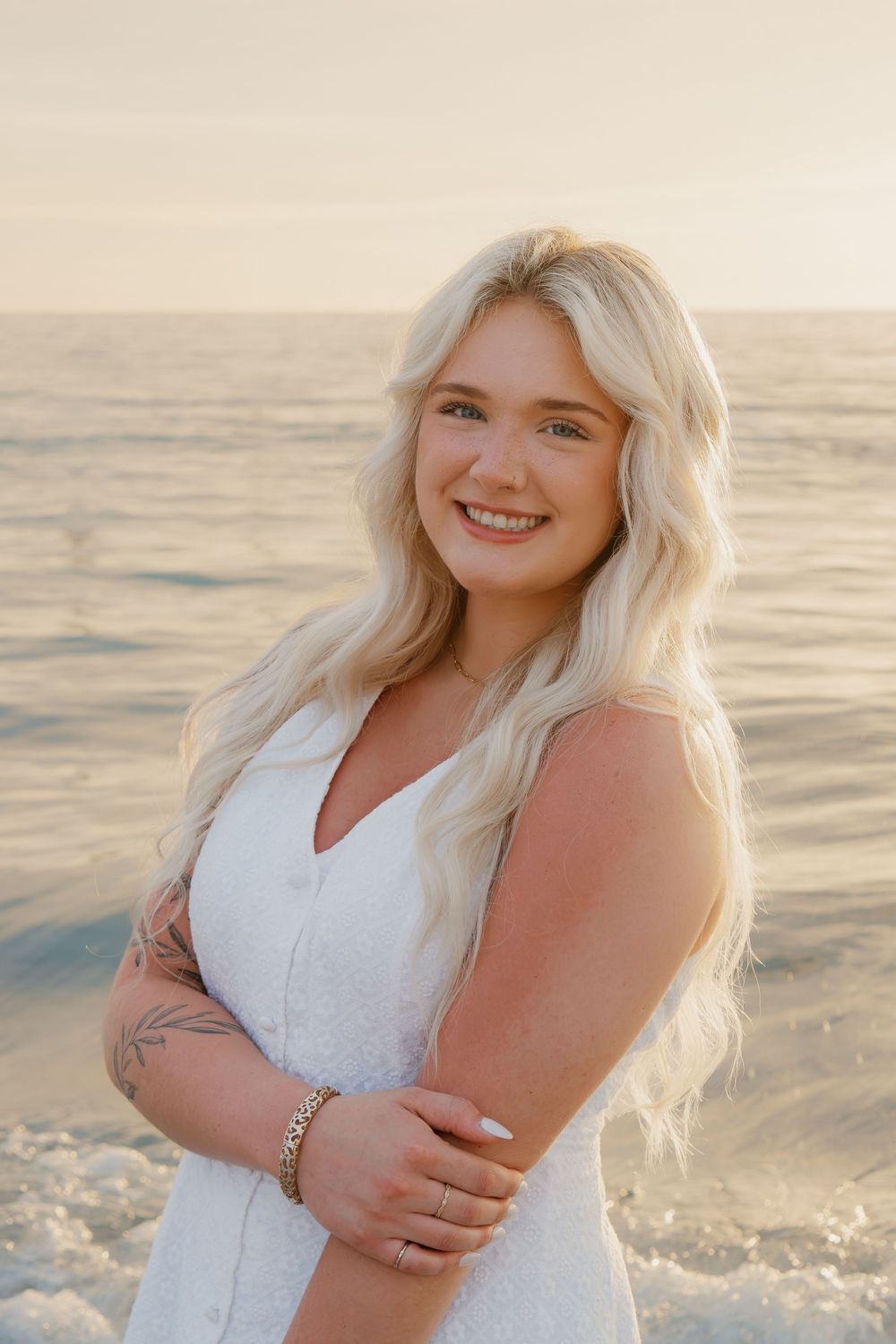 Smiling blonde woman in white dress poses on beach at golden hour with ocean waves in background.