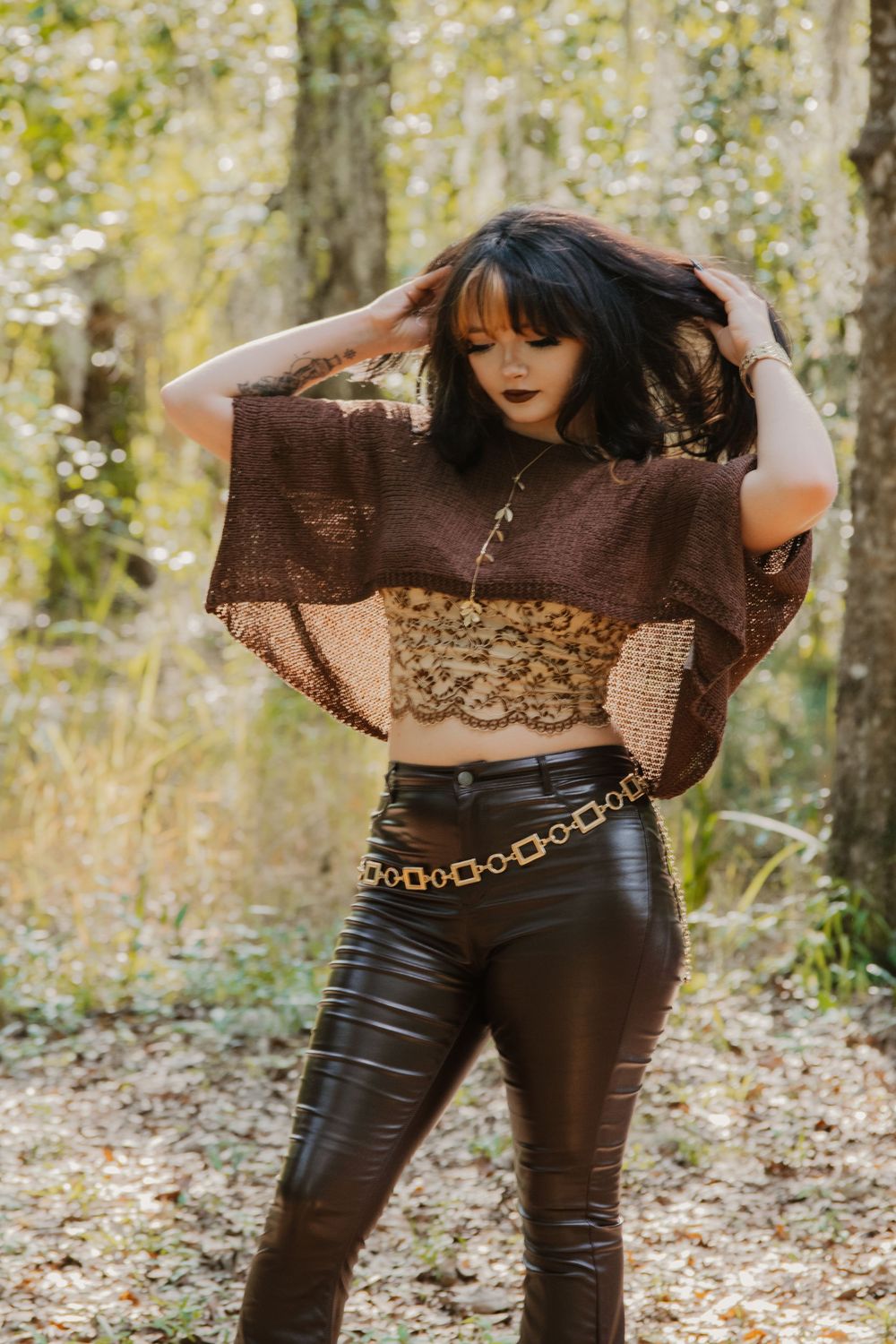 Woman in brown lace crop top and leather pants posing in autumn forest setting.