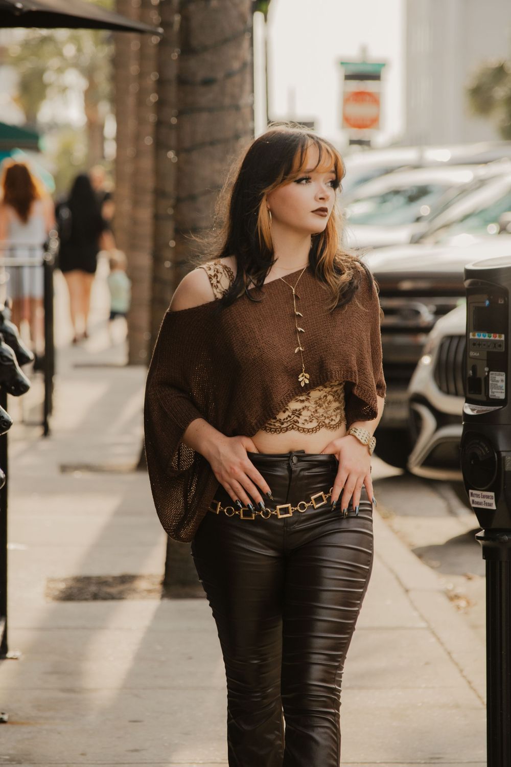 Stylish woman in brown off-shoulder top and leather pants posing on urban city sidewalk.