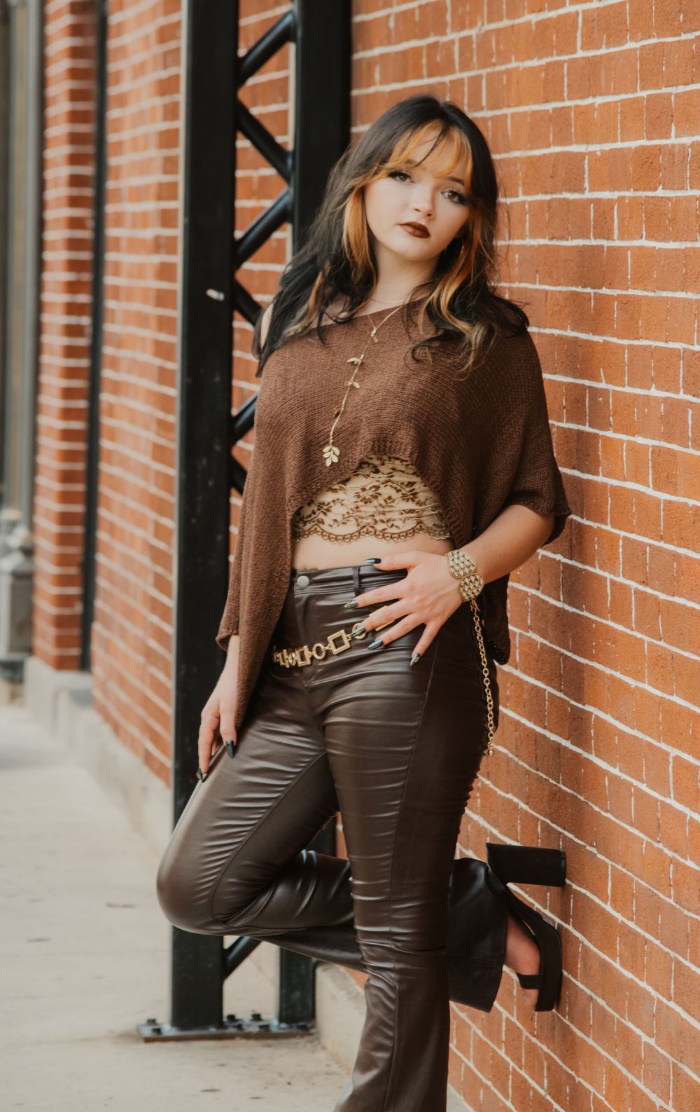 Young woman in brown knit top and leather pants leaning against brick wall in urban setting.