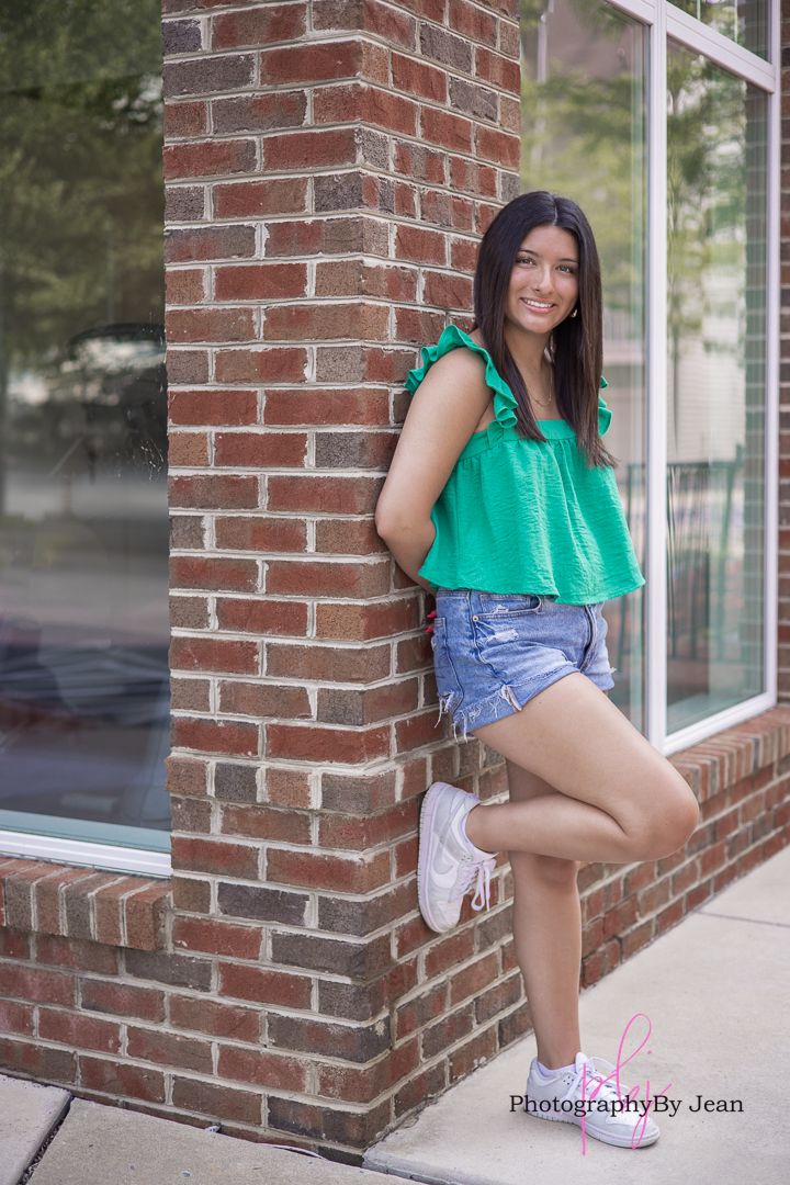 A person in a green sleeveless top and denim shorts poses by a brick wall near a window.