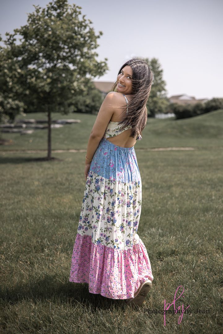 A person in a floral print tiered maxi dress poses outdoors in a grassy park setting on a summer day.
