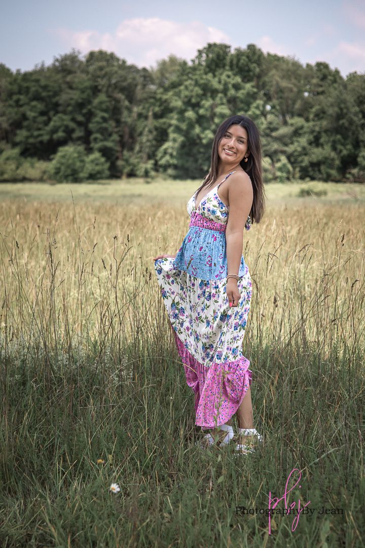 A person in a floral maxi dress poses in a sunlit meadow with tall grass and trees in the background.