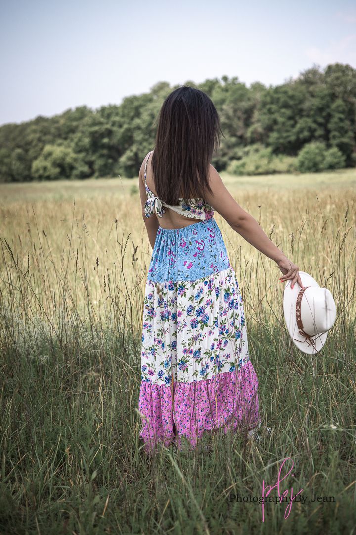 Woman in a floral tiered sundress holds a white sun hat while standing in a summer meadow with trees in background.
