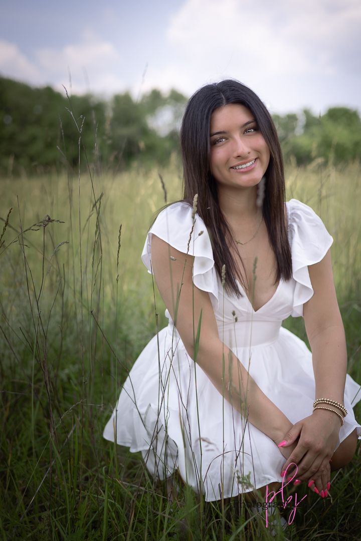 Woman in white ruffled dress sits in tall grass field on a sunny day.