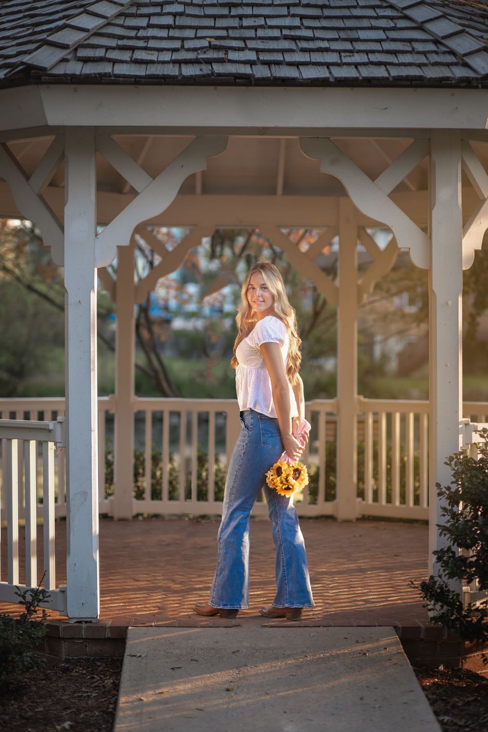 Young woman in flare jeans holding sunflowers smiles inside a white wooden gazebo in golden hour light.