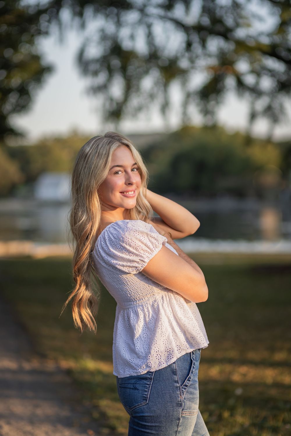 Young blonde woman in white off-shoulder top and jeans smiling outdoors in golden hour sunlight near a park.