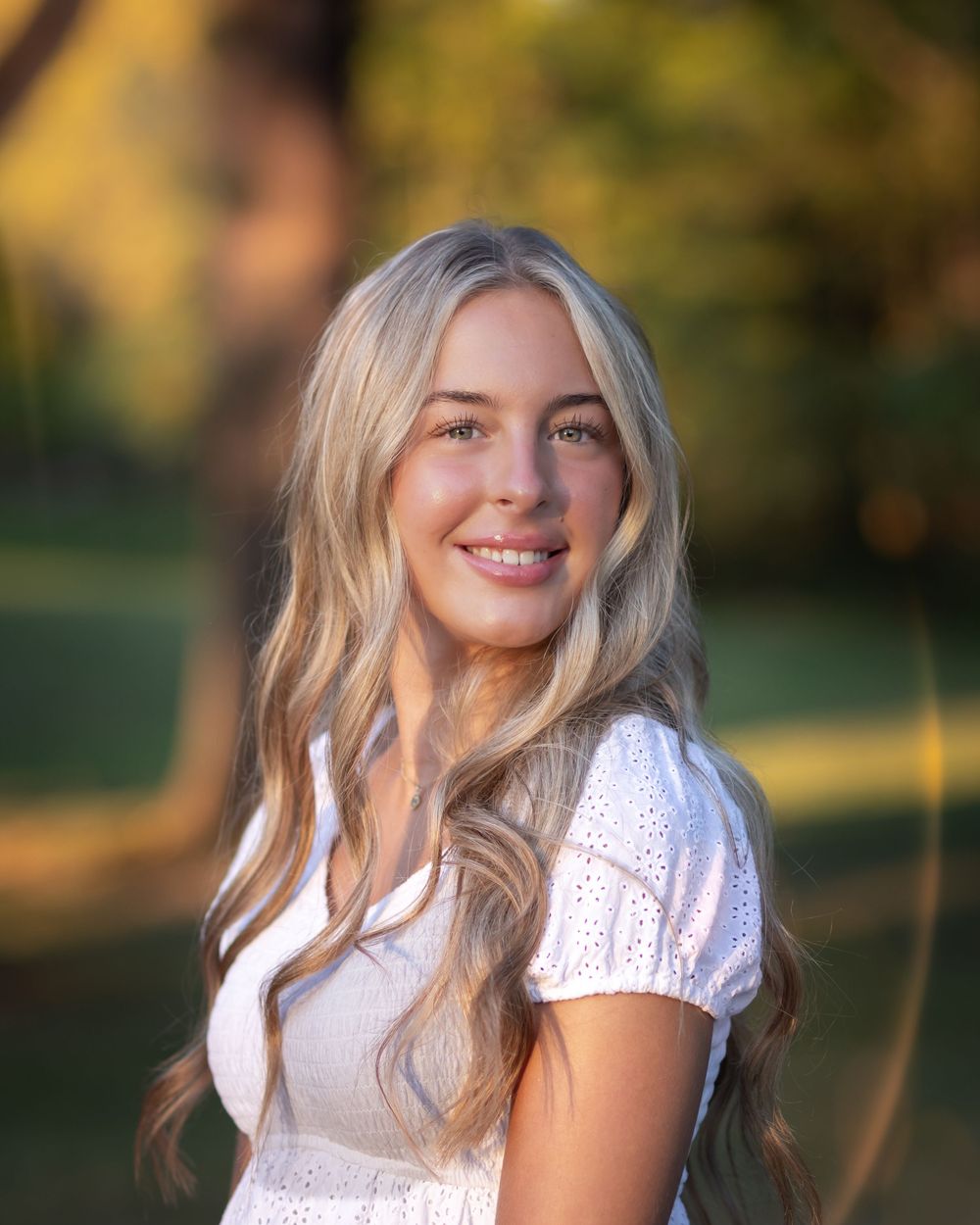 Young blonde woman with long wavy hair smiling outdoors in warm golden sunlight, wearing a white eyelet dress.