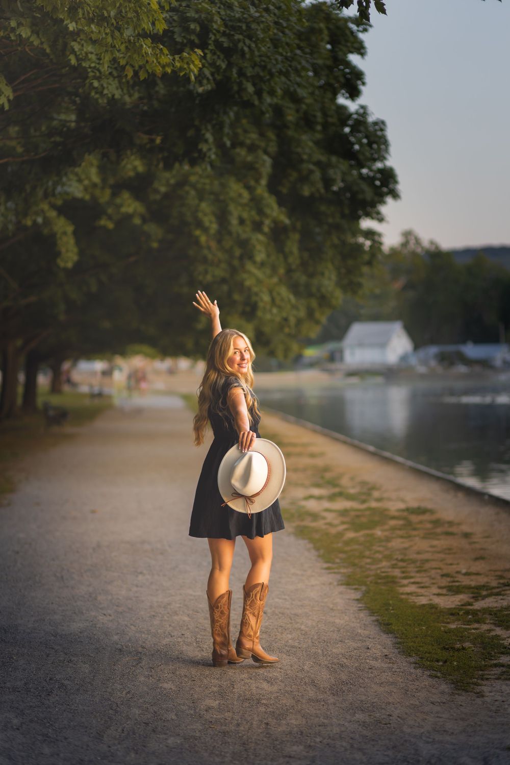 Blonde woman in black dress and cowboy boots holding white hat, waving by a sunlit lakeside path.