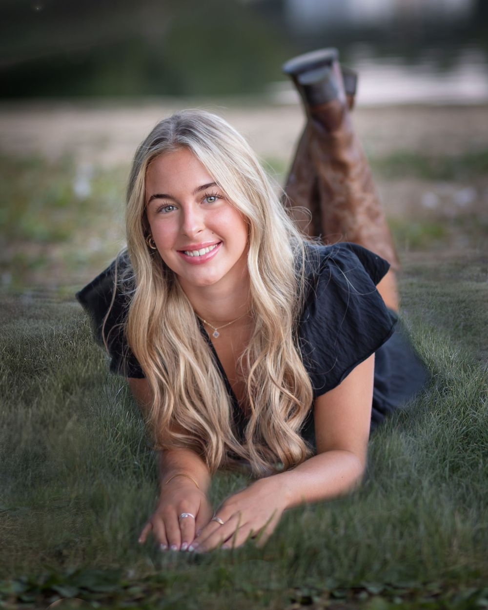 Young blonde woman in black top and boots lying on green grass, smiling at camera in a portrait photo.