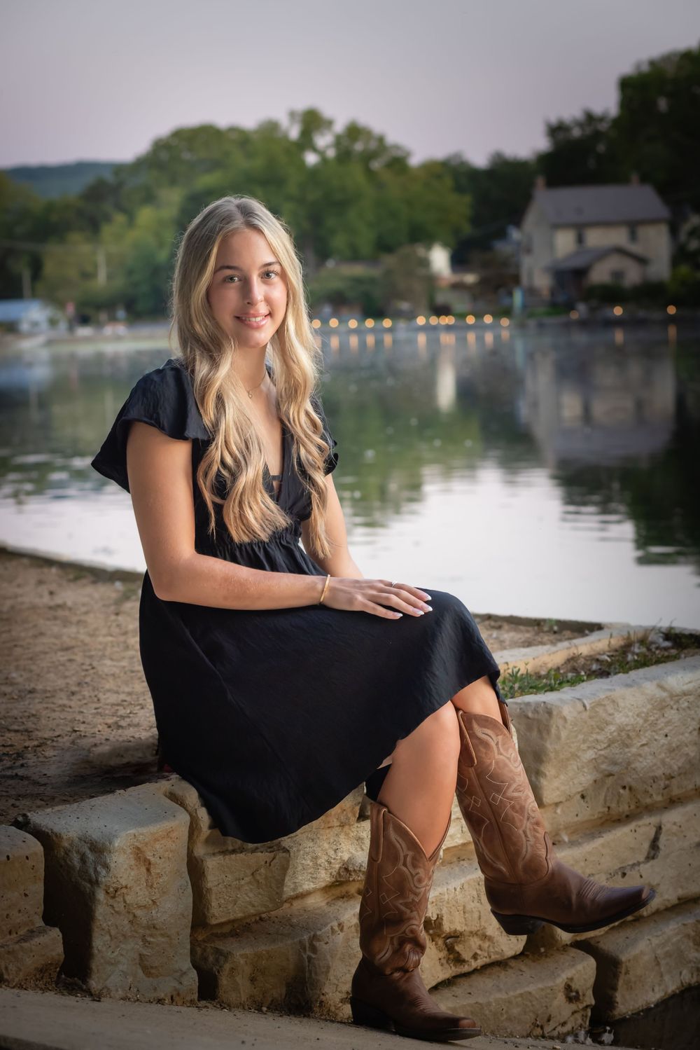 Smiling blonde teen in black dress and cowboy boots sitting on stone steps by a serene lake at dusk.